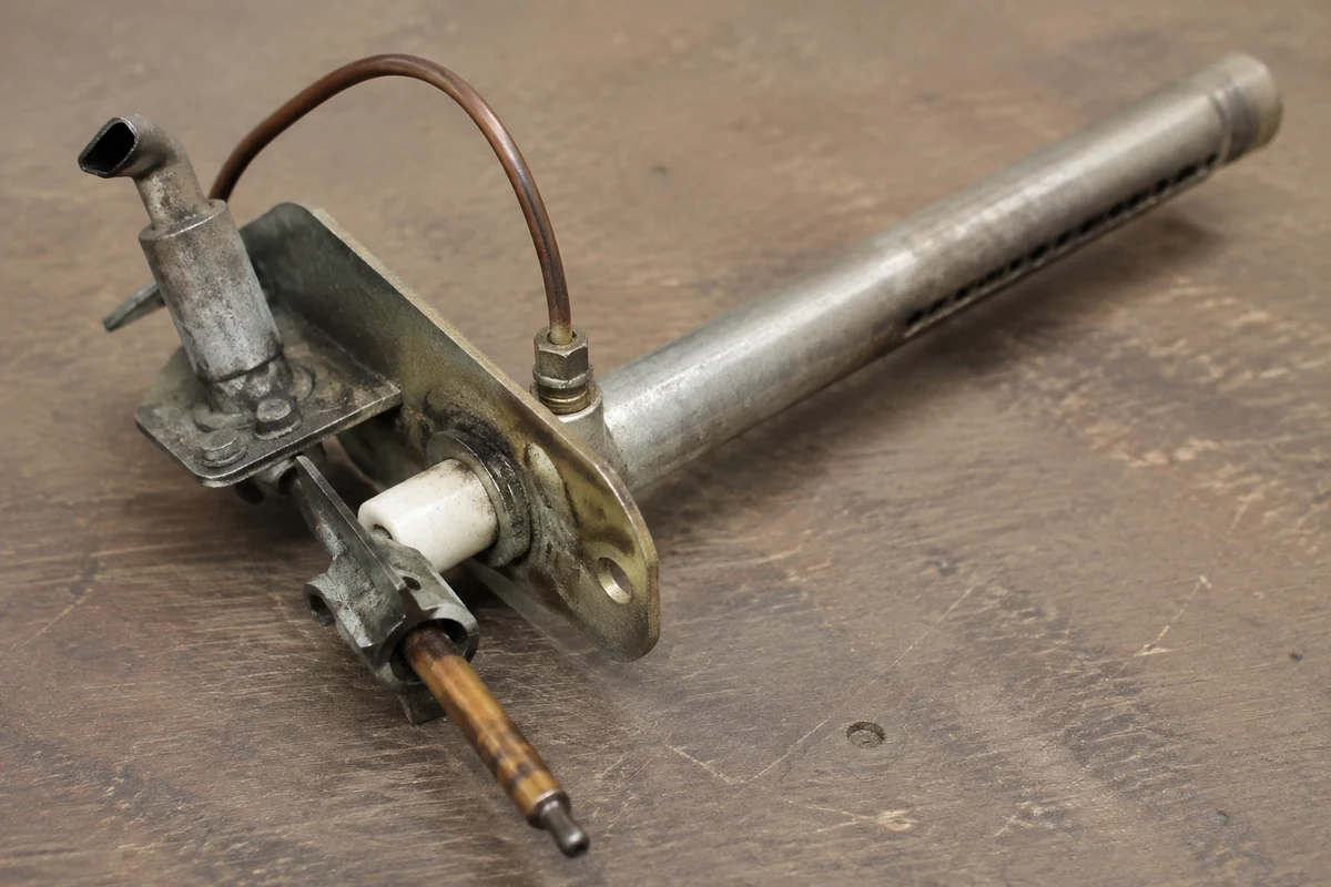 A gas water heater burner assembly on a bench, with the pilot burner, thermocouple and main burner tube