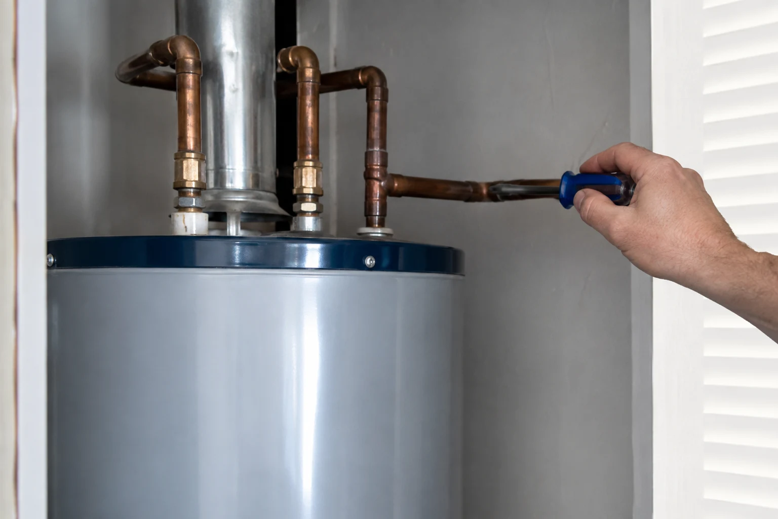 A hand pointing to the hot and cold heat-trap nipples on top of a residential water heater and to the supply pipe where it passes through a wood floor joist, the two spots that produce a benign clicking sound