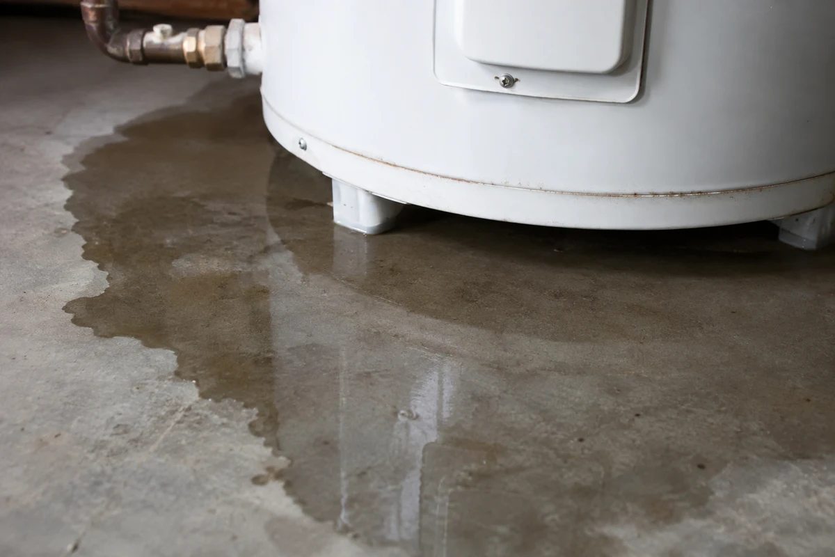 A shallow puddle of water spreading across a concrete floor at the base of a white water heater tank